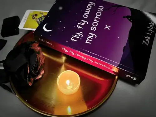 The novel, 'Fly, Fly Away My Sorrow' by Zak Lylak resting on a golden plate near a lit tealight candle, and a black plastic rose. 'The Fool' tarot card is poking from the top of the book.