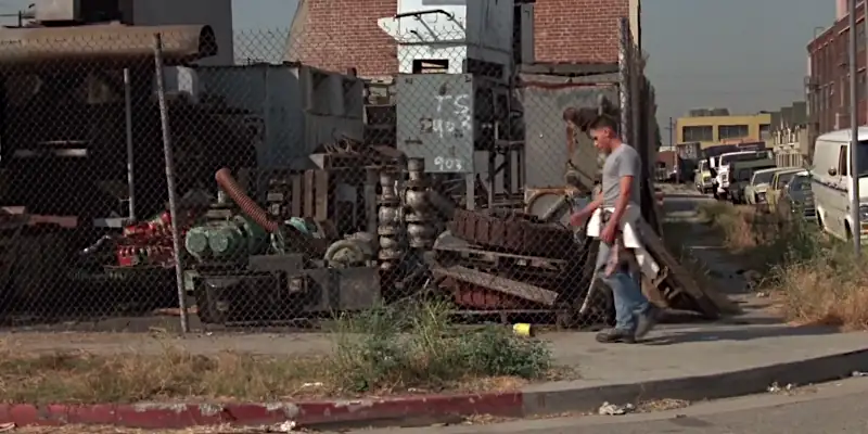 Otto from Repo Man walking along the sidewalk next to a derelict, rusting factory