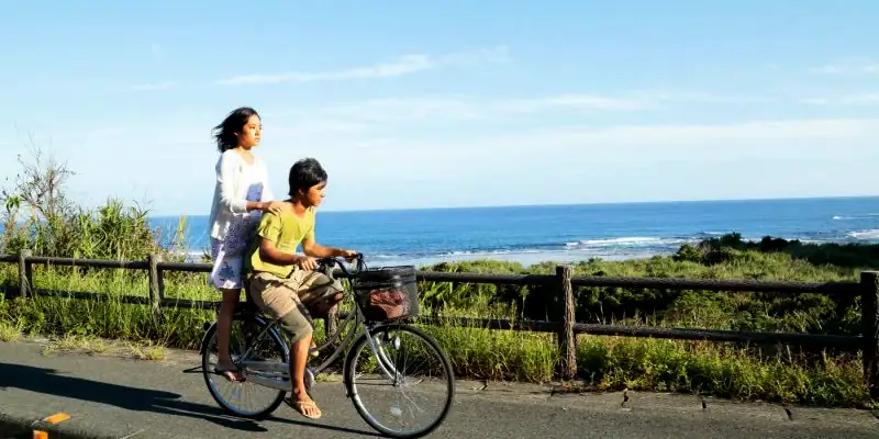 A scene from Still the Water where Kaito and Kyoko are cycling down a road near the sea